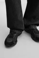 Zapatilla deportiva tipo running de color negro, perteneciente a la colección especial Origins. Presenta una parte superior confeccionada en una combinación de cuero con acabado de gamuza y tejido transpirable. Cuenta con una suela de volumen con diseño irregular y cierre mediante cordones.