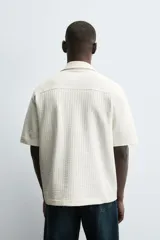 Camisa de manga corta con cuello bowling y calce relaxed fit, confeccionada en tejido seersucker elástico con textura. Presenta un diseño a rayas verticales en tonos oscuros y cierre frontal con botones.