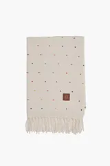 Pashmina de color crudo con estampado de pequeños puntos multicolores y flecos en los extremos. Cuenta con una etiqueta de cuero sintético con logo grabado.