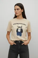 Remera de algodón color beige, de corte holgado y cuello redondo, con estampado frontal de un oso con lentes de sol y la frase 'slaying everyday'.