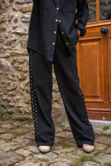 Pantalón de denim negro de corte amplio, con cierre de botón y cremallera. Presenta detalles de tachas doradas en los laterales y bolsillos.