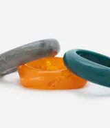 Set de tres brazaletes rígidos de resina en diferentes colores: gris marmolado, naranja traslúcido y verde esmeralda. El brazalete naranja tiene forma cuadrada con bordes redondeados.