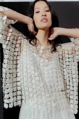 Blusa de red en color blanco con apliques de flores tridimensionales en toda la prenda. Presenta transparencias, cuello redondo y mangas largas.