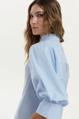 Camisa celeste de manga larga abullonada, con cuello mao decorado con un pequeño volado y puños anchos. Confeccionada en mezcla de algodón, poliamida y elastano.