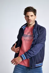 Campera liviana de nylon color gris oscuro, con cierre frontal completo, cuello subido y puños y cintura acanalados con rayas en rojo y blanco. Presenta logo bordado en el pecho y bolsillos laterales con cierre.