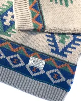 Buzo tejido de punto con diseño geométrico inspirado en motivos andinos, en tonos beige, verde, azul y naranja. Presenta cuello redondo, puños y ruedo acanalados, y etiqueta de marca en el frente.