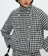 Saco corto estructurado con estampado pied de poule en blanco y negro. Presenta cuello alto, cierre frontal cruzado con botones decorativos y mangas largas.