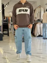 Hoodie de corte clásico con medio cierre y cuello alto, en color marrón con una franja horizontal blanca en el pecho que incluye el logo estampado en negro. Presenta además un pequeño bordado en el hombro izquierdo y puños y cintura elastizados de rib.