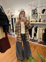 Camisa de corte relajado con estampado de cuadros tartán en tonos marrones y beige. Presenta cuello clásico, cierre frontal con botones, mangas largas con puños abotonados y bolsillos frontales en el pecho.