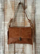 Cartera bandolera de cuero color tostado, con solapa frontal, cierre metálico de gancho y tiras decorativas nudosas en las asas.