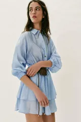 Vestido corto de color celeste con cuello solapa, cierre frontal de botones y manga larga con puños elásticos. Presenta cintura elástica, falda con volantes y un cinturón fino con un pequeño bolsillo de flecos incorporado.
