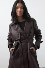 Trench coat de símil cuero color marrón oscuro, con solapas amplias, cierre frontal con botones, cinturón ajustable en la cintura y mangas con tiras ajustables. Presenta un largo por debajo de la rodilla y bolsillos laterales.