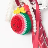 Llavero decorativo con mosquetón metálico en forma de corazón, que incluye una variedad de elementos colgantes: una pieza tejida a crochet con forma de sandía, una tira de perlas, cintas de raso rojas y cordones deportivos con punteras metálicas.