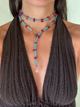 Collar largo estilo lariat, compuesto por bastones de vidrio facetados color celeste/azul claro y cuentas irregulares de piedra color coral/rojo. Cuenta con herrajes de acero quirúrgico y un dije colgante con un logo grabado.