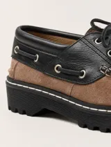 Mocasín de cuero con diseño bicolor en negro y marrón, con detalle de flecos sobre el empeine, cordones y suela track cosida.