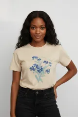 Remera de algodón color beige con estampado floral en tonos azules en el centro del pecho y detalle de texto manuscrito.