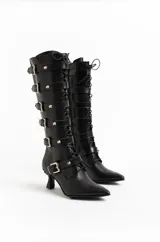 Botas altas de cuero negro con acabado brillante texturado, múltiples tiras con hebillas y tachas plateadas, cordones frontales, punta fina y taco bajo escultural.