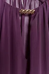 Blusa de tejido semitransparente color violeta, con cuello redondo y detalle de fornitura dorada en el escote. Presenta un diseño de escote en pico, mangas largas abullonadas con puños elásticos y corte holgado.