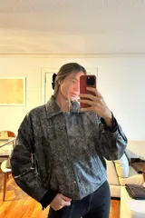 Campera bomber de cuero ovino con efecto desgastado en tonos grisáceos y negros. Presenta cuello camisero, cierre frontal con broches forrados y diseño holgado.
