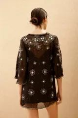 Vestido corto negro de tela transparente con mangas acampanadas y escote en V. Está completamente bordado con motivos florales y estrellas en lentejuelas y cuentas plateadas y verdes.