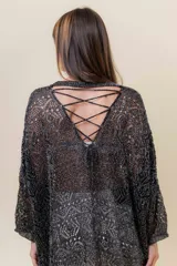 Buzo de tejido liviano y calado con brillo sutil, color gris oscuro o negro. Presenta un calce amplio y mangas amplias, con un detalle de tiras entrelazadas en la espalda. El largo es irregular, más largo en la parte trasera.