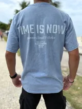 Remera oversize de color celeste melange, con diseño de estampado tipográfico en la espalda que incluye la frase "Time is Now" y otros detalles de colección.