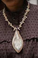 Collar corto con dije central en forma de hoja, elaborado en guampa con engarce de alpaca. La cadena está compuesta por bastoncitos de acrílico y mostacillas transparentes, finalizando con una cadena de eslabones de acero quirúrgico ajustable.