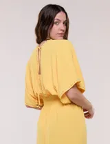 Vestido largo amarillo de corte fluido con escote en V, mangas cortas amplias y apliques de tachas en el escote y puños. Posee cintura elastizada fruncida y falda larga con tajos laterales en el ruedo.