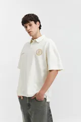 Remera polo color crema con logo bordado en el pecho, de corte amplio y mangas cortas.