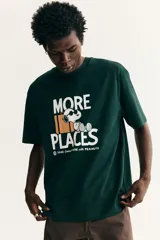 Remera verde oscuro con estampado frontal de Snoopy, equipaje y la inscripción "More Places - Take (more) care with Peanuts". Cuenta con cuello redondo y mangas cortas con hombros caídos.