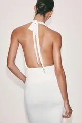Vestido midi blanco de cuello halter, confeccionado con mezcla de poliamida. Presenta detalle de drapeado lateral con un broche metálico dorado en forma de caracola y cierre en la espalda ajustable con lazadas.