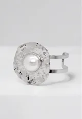 Brazalete rígido de diseño circular con acabado plateado texturizado e irregular, destacando una perla central grande. Presenta un estilo moderno y llamativo.