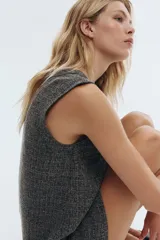 Vestido corto sin mangas, de corte recto, con cuello redondo y cierre frontal de botones. El tejido es tweed o similar, en tonos grises y negros jaspeados.