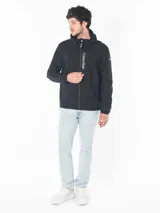 Campera rompeviento Calvin Klein de color negro, con cierre frontal, capucha ajustable y logo estampado verticalmente en el frente. Presenta puños elásticos y un parche con el logo en la manga.