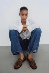 Mocasín de cuero marrón con textura granulada, diseño de horma cuadrada, detalle de flecos y lazo decorativo en el empeine, montado sobre una suela y taco bajo de color negro.