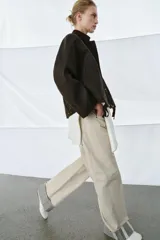 Chaqueta encerada de corte oversize, color marrón oscuro, con cuello de pana en contraste. Presenta cierre frontal con cremallera y botones a presión ocultos bajo solapa, bolsillos delanteros y mangas largas con puños ajustables.