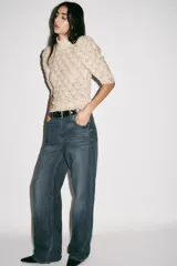 Jersey de punto abierto color crema, con cuello subido y mangas cortas abullonadas. Presenta un patrón de tejido calado con motivos en forma de hoja o escama y un borde festoneado en el ruedo.