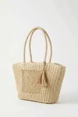 Cartera tipo shopper de mimbre tejido color beige, con doble asa de hombro y detalle de borla colgante.