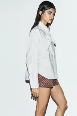 Camisa de popelín de algodón con cuello subido y detalle de aplicación tipo pañuelo anudado al cuello. Presenta manga larga con puños clásicos y cierre frontal mediante botones.