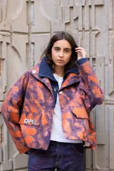 Campera de corte oversize con estampado abstracto en tonos naranja y violeta. Presenta cuello alto tejido en color azul oscuro, cierre frontal con botones metálicos a presión y bolsillos frontales con solapa, uno de ellos con el detalle de la palabra 'CAPER' aplicada.