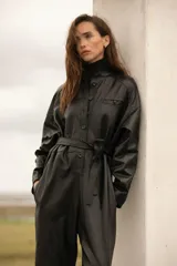 Mameluco negro de cuero ovino, con cuello alto, cierre frontal con botones a presión, cinturón con hebilla forrada y bolsillos laterales.