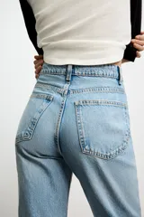 Jeans de mujer, tiro alto, color azul medio con efecto desgastado (lavado claro en muslos y oscuro en bordes). Corte recto y ancho (mom fit) con cinco bolsillos y cierre frontal de botón y cremallera.