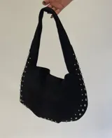 Cartera tipo hobo de gamuza negra, con diseño slouchy y tachas metálicas plateadas decorativas a lo largo de los bordes y la base. Cuenta con un asa integrada para llevar al hombro y un bolsillo interno con cierre.