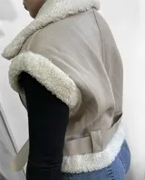 Chaleco color beige con interior de piel sintética blanca, cierre frontal con cremallera y cinturón ajustable en la cintura.