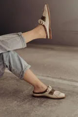 Zueco de cuero color crudo, con diseño destalonado, punta cuadrada, detalle de flecos y una pieza central de cuero con estampado snake en tonos marrones y un aplique metálico triangular.