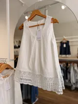 Blusa blanca sin mangas, confeccionada en mezcla de lyocell, nailon y lino, con escote redondo, pliegues frontales y detalle de puntilla en el ruedo.