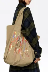 Bolso tipo shopper de color beige, con diseño amplio y doble asa de hombro. Presenta un detalle frontal de bordados coloridos con hilos colgantes que forman un diseño artístico.