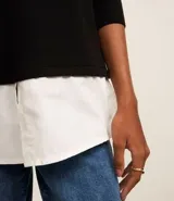 Suéter negro de punto con cuello redondo y mangas largas. Tiene un dobladillo superpuesto de camisa blanca.