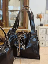 Bolso tipo shopper de cuero con acabado brillante efecto craquelado en color negro. Presenta correas de hombro integradas, cierre con cordones ajustables en el frente y detalles de costuras visibles en los bordes.