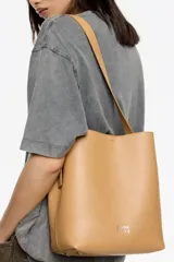 Bolso de hombro Bimba y Lola modelo Carlino, de estructura rígida y tamaño mediano, confeccionado en piel vacuna color tostado. Presenta asa regulable de piel con hebilla metálica y logo impreso en la parte inferior.